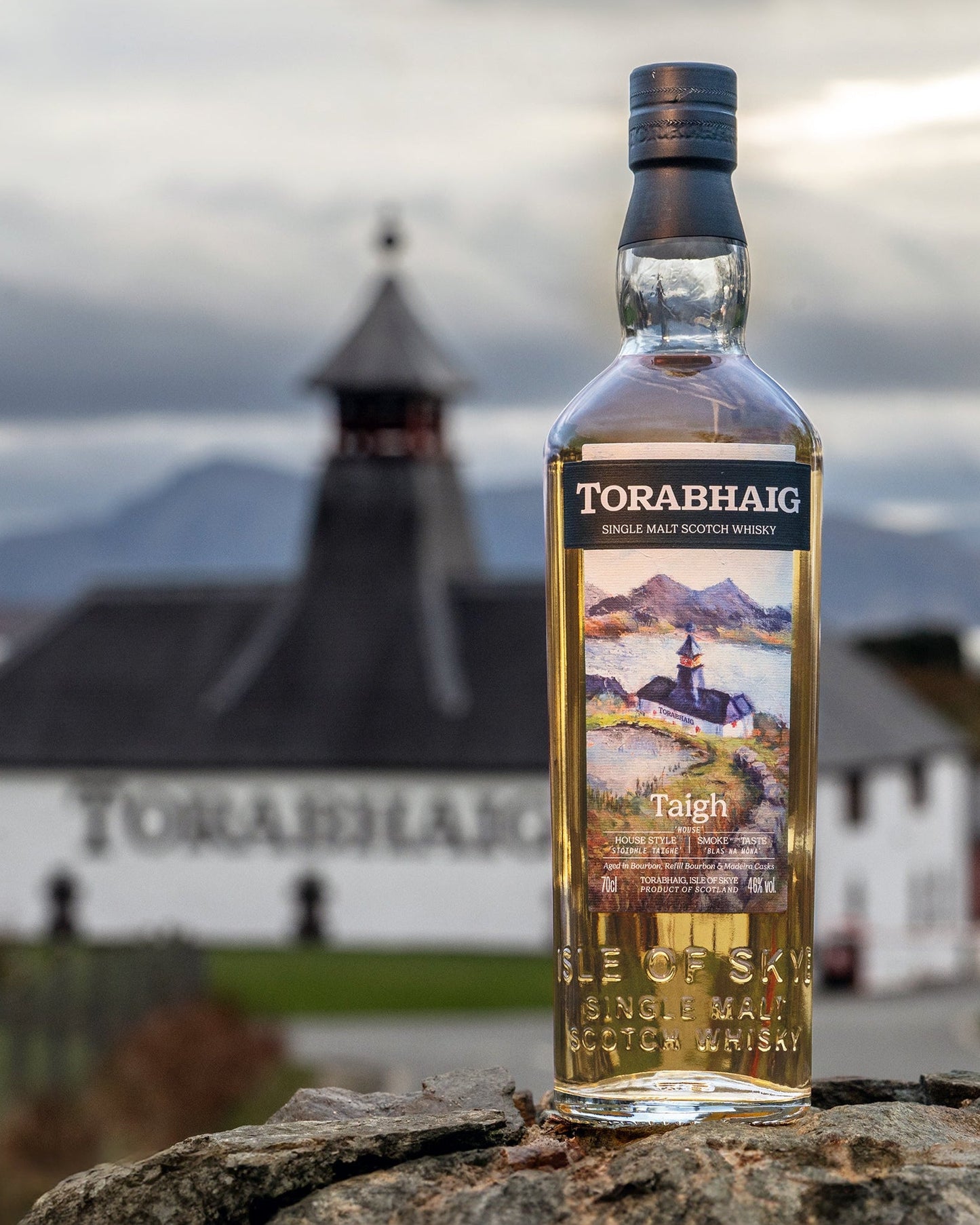 Torabhaig whisky bottle with a scenic label against a blurred background of a building and mountains.